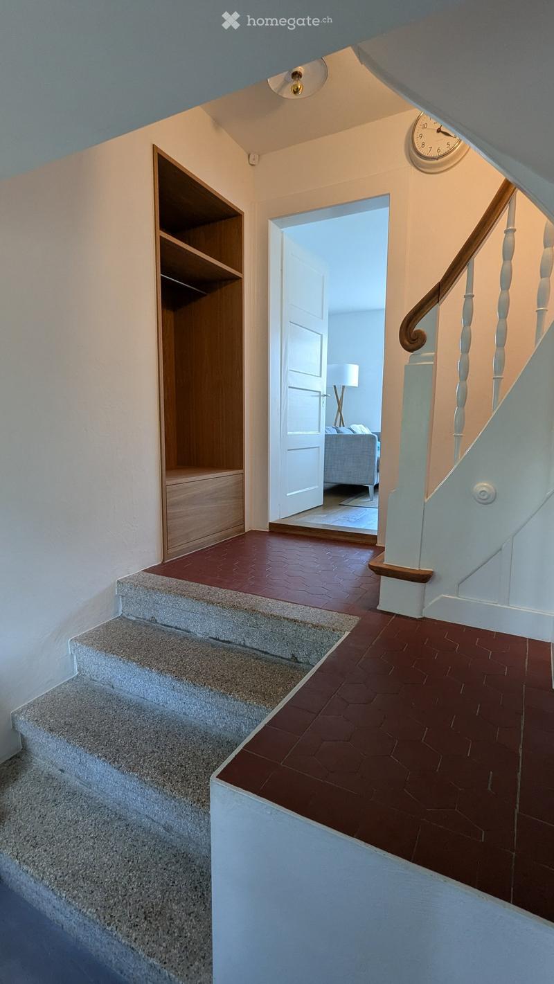 Staircase with red hexagonal tiles, white walls, wooden handrail, and a small built-in shelf on the left side. A doorway leads to another room with a visible sofa and lamp.