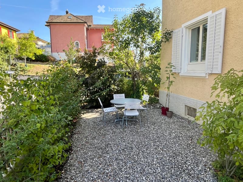 A small, well-maintained garden area with a round table and four chairs, surrounded by gravel and lush greenery, with a building in the background.