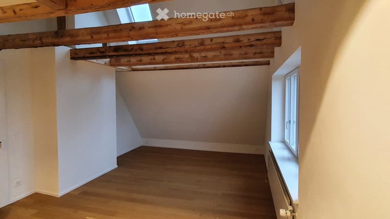 Empty attic room with wooden beams on the ceiling, wooden flooring, and a single window with white frame.