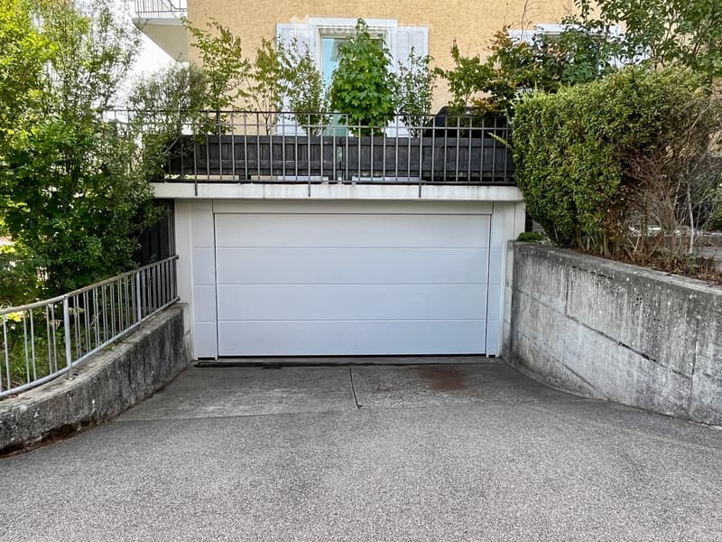 A modern building entrance with a large garage door, metal railing, and greenery surrounding the area.