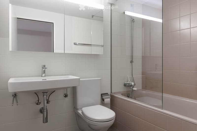 Modern bathroom with white tiles, a sink, toilet, bathtub with glass shower enclosure, and a mirror above the sink.