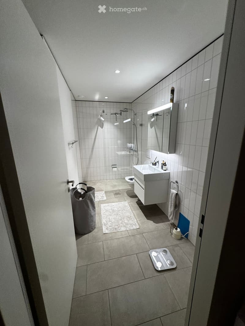Modern bathroom with white tiles, a sink with a mirror, a toilet, a shower, and a towel hanging on a rack. A scale and a handbag are also visible.