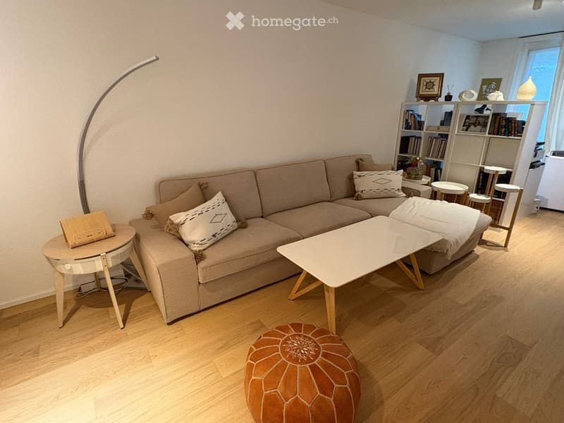 Modern living room with a beige sofa, coffee table, ottoman, side table, bookshelf, and floor lamp. The room features wooden flooring and large windows with white curtains.