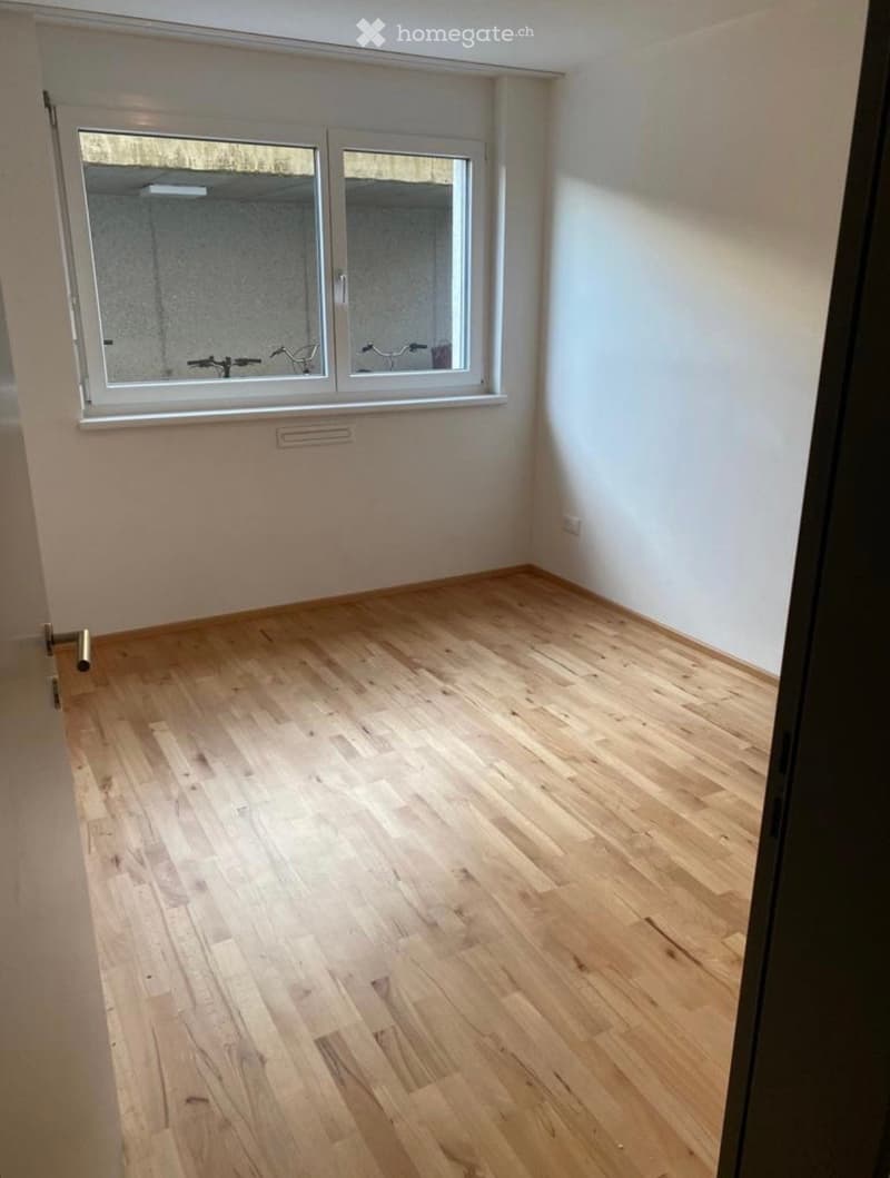Empty room with light wooden flooring and white walls, featuring a large window with a view of bicycles outside.