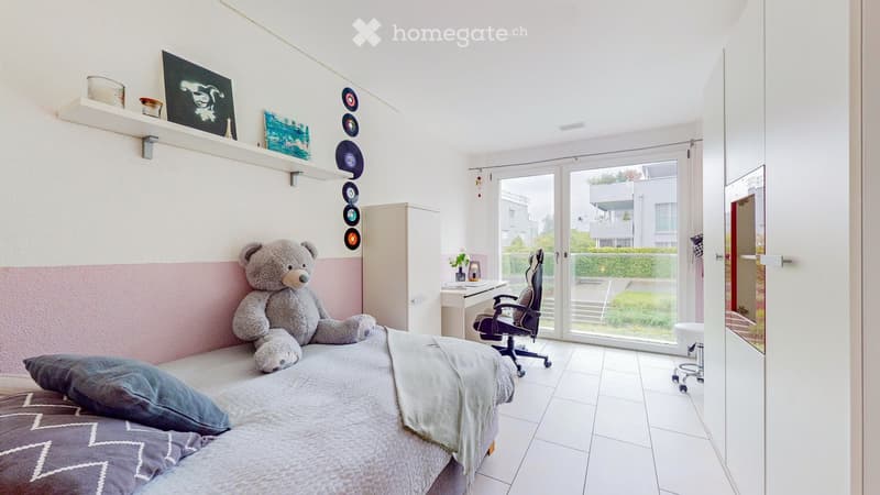 A modern bedroom with a bed, pillows, a blanket, a large teddy bear, a desk with a chair, a cabinet, a shelf with picture frames and vinyl records, and a large window with a view of the garden.