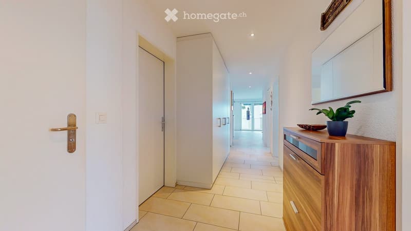 Bright hallway with tiled floor, white walls, and a small wooden console table with a potted plant and decorative bowl on top. A closed door with a brass handle is visible on the left.