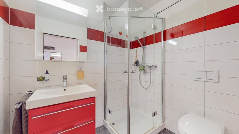 Modern bathroom with white tiles, red accents, a glass-enclosed shower, a sink with a red cabinet, and a toilet.