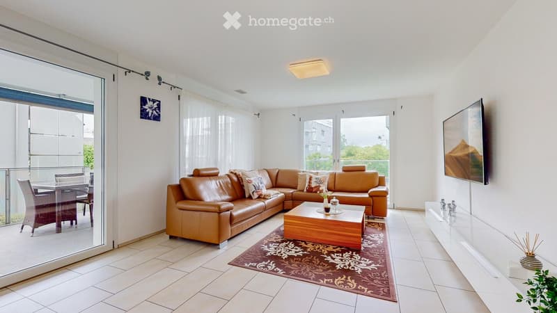 Bright living room with a large leather sectional sofa, coffee table, dining area with chairs and table, flat-screen TV, and decorative items including a vase and plant.