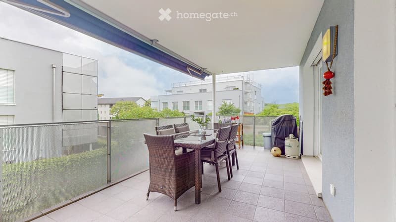 Balcony with a glass railing, tiled floor, and a wicker dining set including a table and chairs. There is a small plant, a decorative ball, and a gas tank visible.