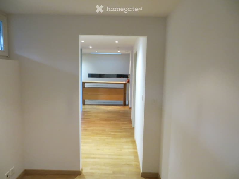 Empty hallway with light wooden flooring and white walls, leading to another room with a visible cabinet and countertop.