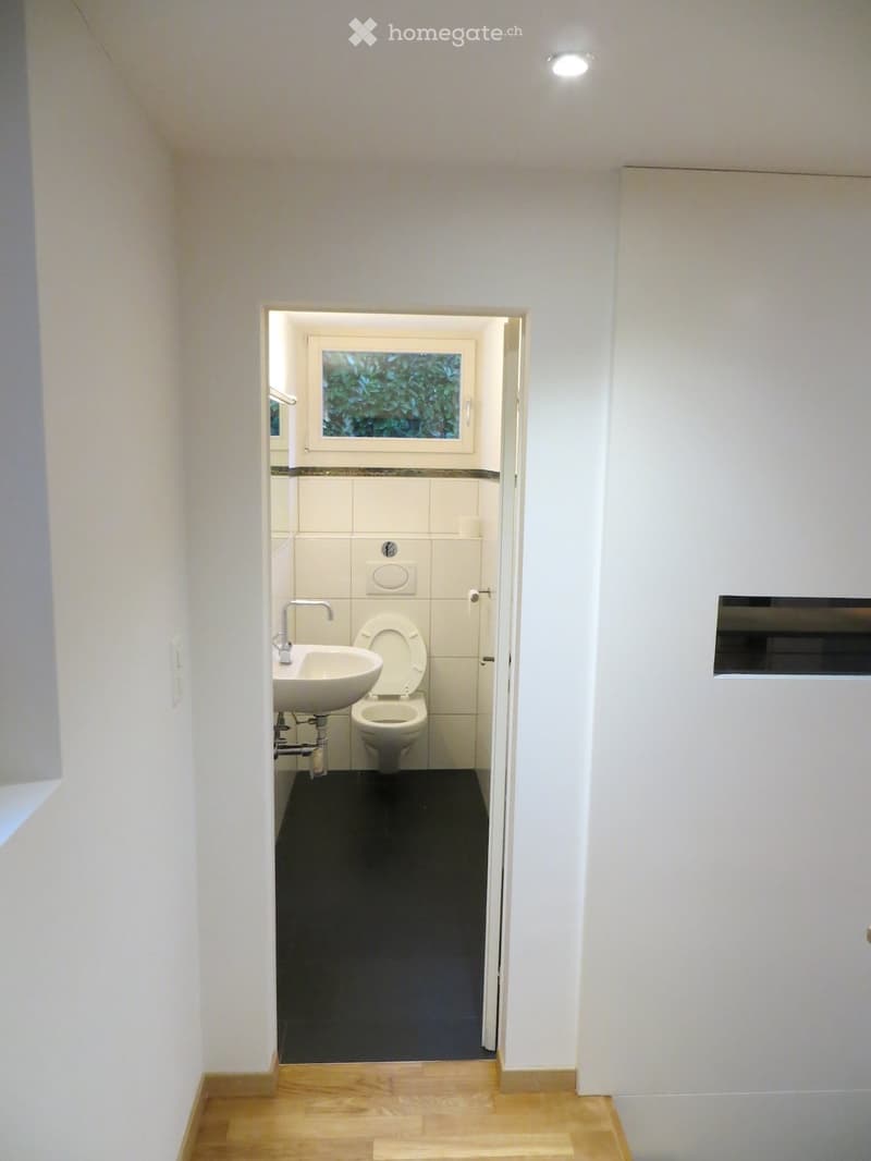 Modern bathroom with white walls, black tiled floor, sink, toilet, and a window with greenery view.