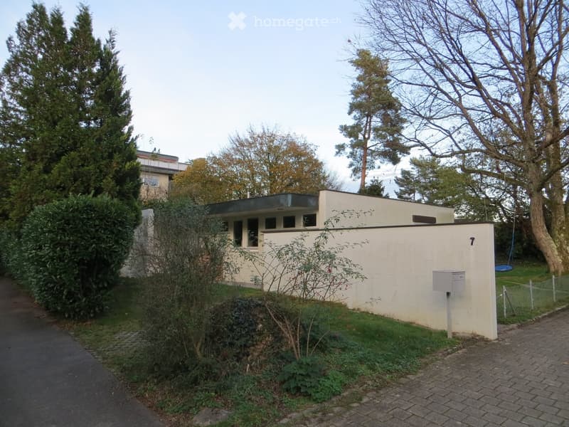 A modern, single-story house with a flat roof and white exterior, surrounded by trees and greenery. The house has a small garden area with a mailbox and a swing visible in the background.