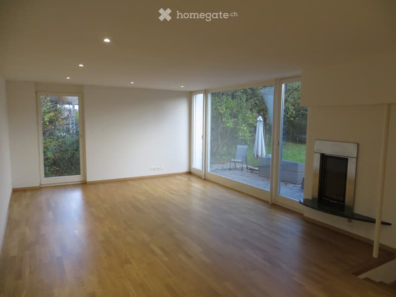 Empty living room with wooden flooring, large windows, and a fireplace. No furniture or equipment present.