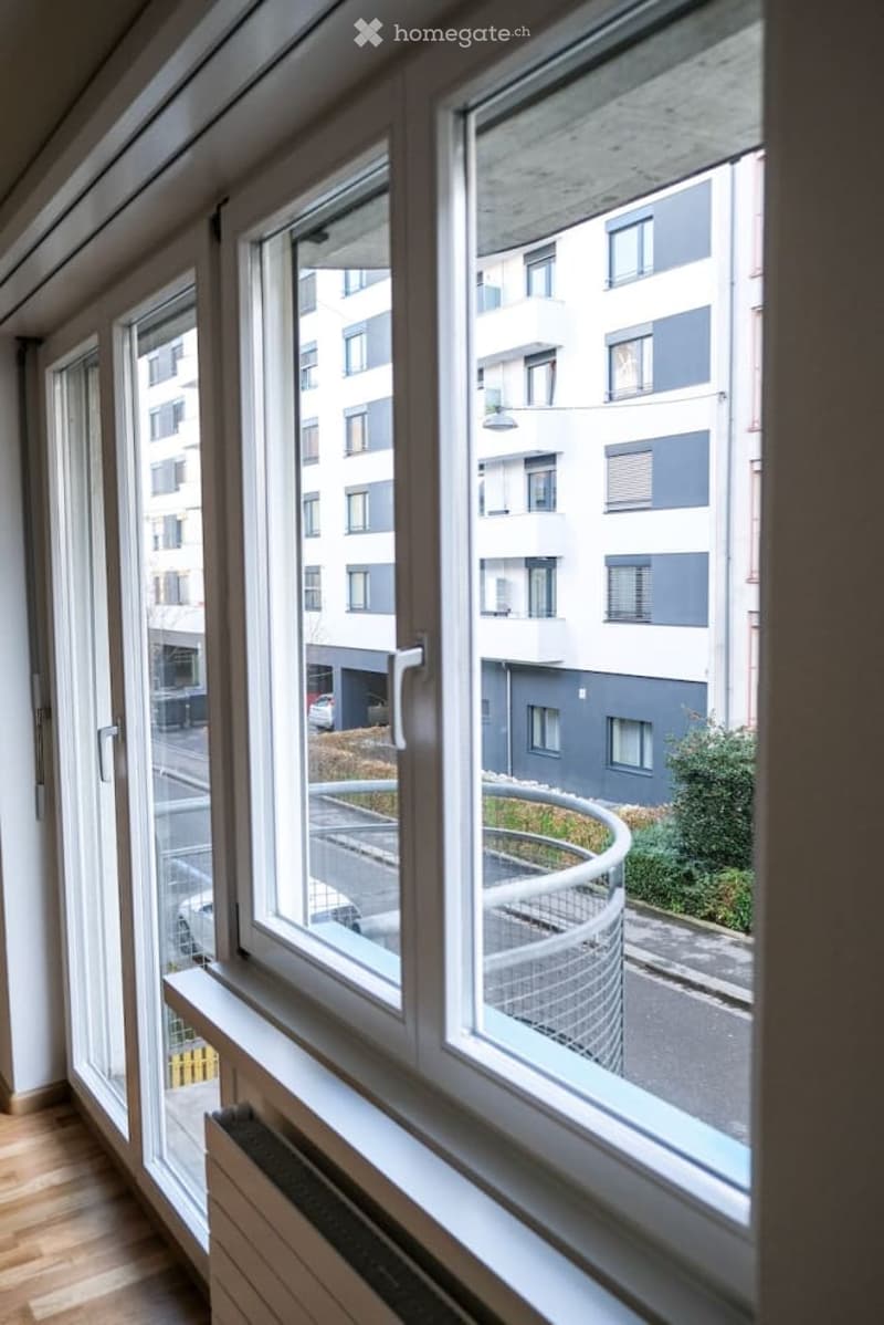 View from a balcony or terrace showing modern apartment buildings, a curved metal railing, and a paved area below.