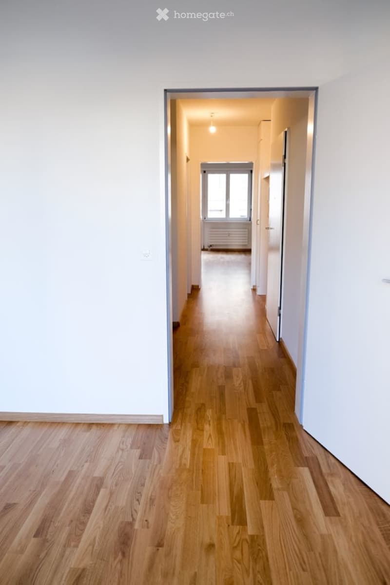 Empty hallway with light wooden flooring, white walls, and a view into an adjacent room with a window and radiator.