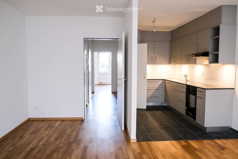 Modern, minimalist kitchen with grey cabinets, white countertops, and dark tiled flooring. The adjacent hallway features light wooden flooring and white walls.