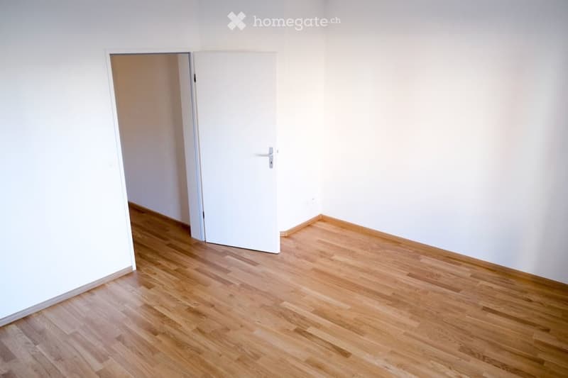 Empty room with light wooden flooring and white walls, featuring a closed white door leading to another space.