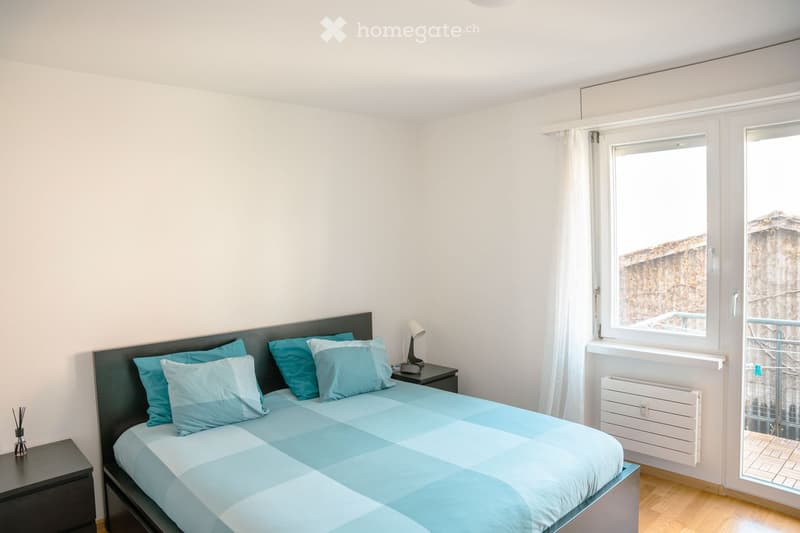 A modern bedroom with a neatly made bed, matching nightstands, and a view through a large window.