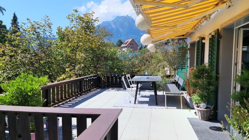 Balcony with a table, chairs, bench, and potted plants, overlooking a scenic mountain view.