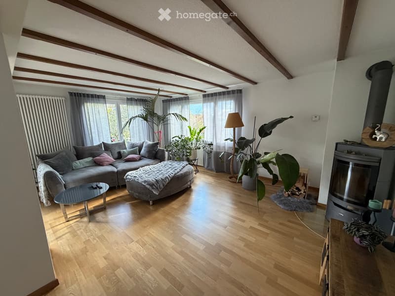 Bright living room with wooden flooring, exposed wooden beams, large windows with curtains, grey sectional sofa, coffee table, floor lamp, fireplace, and potted plants.