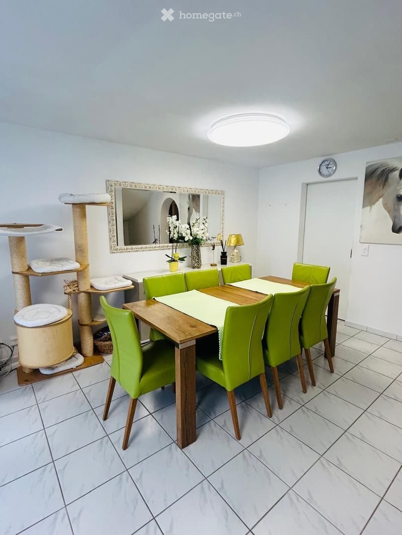 Dining room with a wooden dining table surrounded by green chairs, a cat tree, a mirror, a vase with flowers, and a lamp on the sideboard.