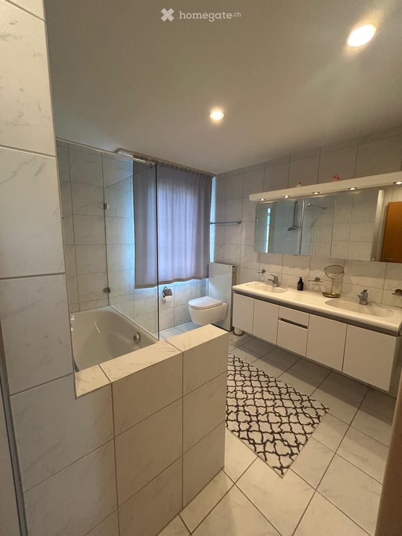 Modern bathroom with a bathtub, toilet, double sink, mirrored cabinet, and a large window with a curtain. The space features tiled walls and floors, recessed lighting, and a decorative black and white rug.
