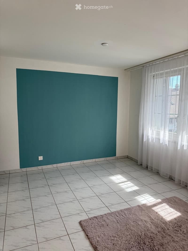 Empty room with tiled floor, white walls, large window with sheer curtains, and a single area rug.