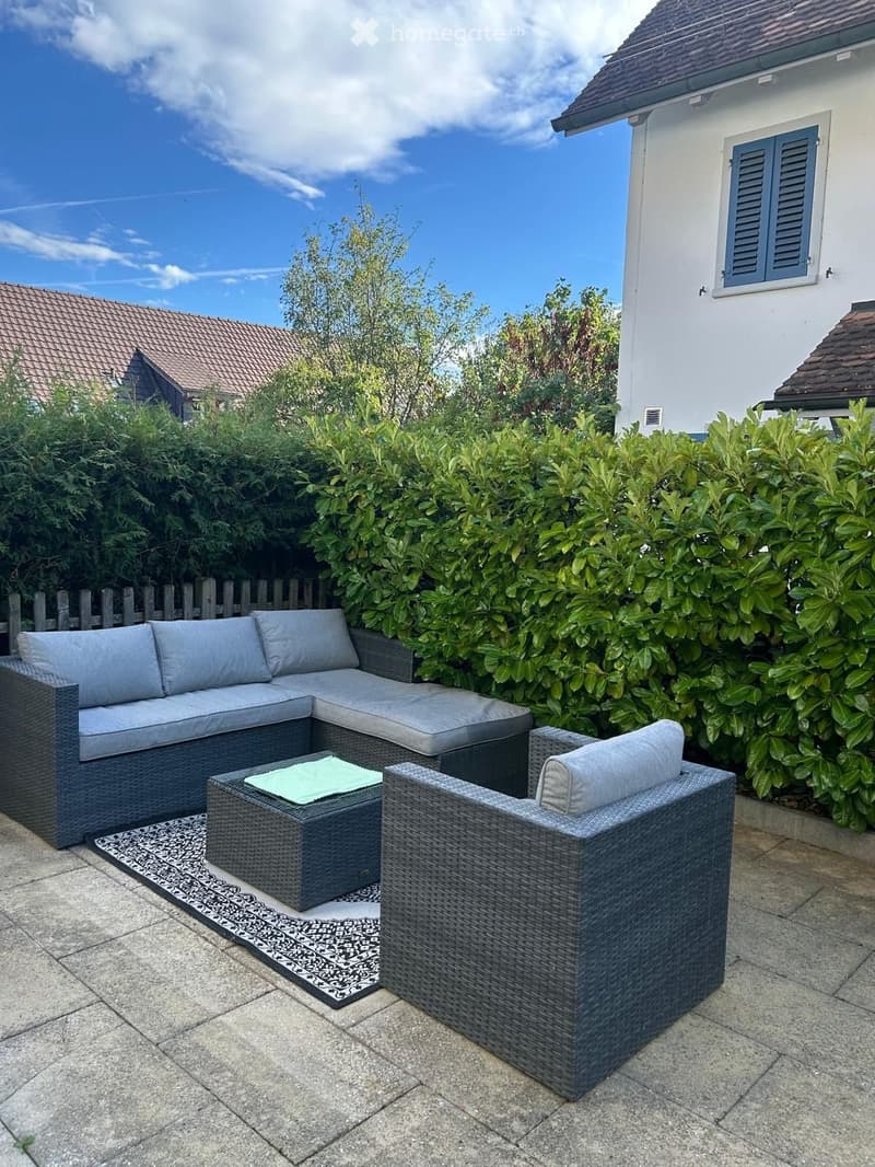 A patio area with a grey wicker sofa, matching chair, and a small table with a green cushion, surrounded by lush greenery and a wooden fence.