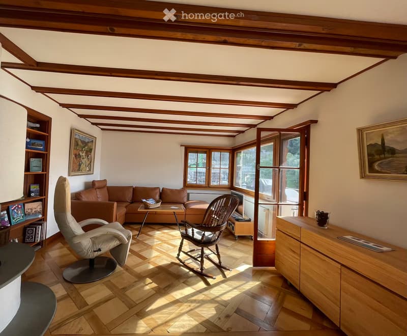 Cozy living room with wooden beams, hardwood floor, large windows, brown leather sofa, coffee table, rocking chair, and bookshelf.
