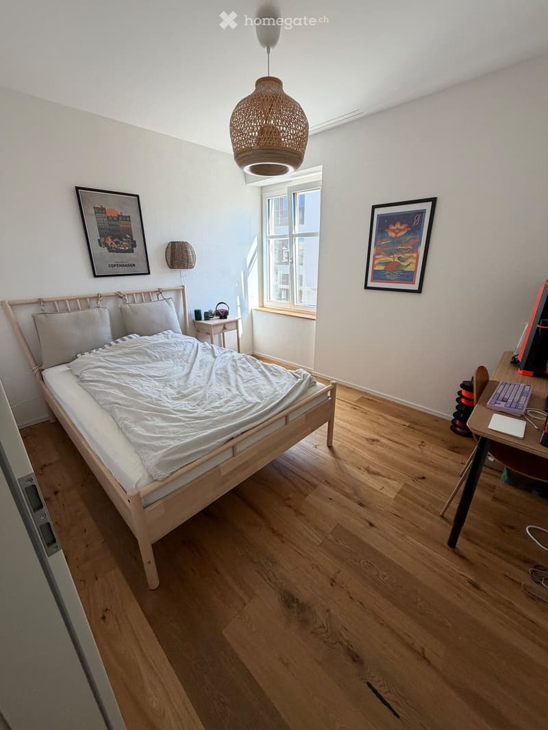 Cozy bedroom with wooden floor, single bed, desk with laptop and keyboard, nightstand, and framed artwork on walls.
