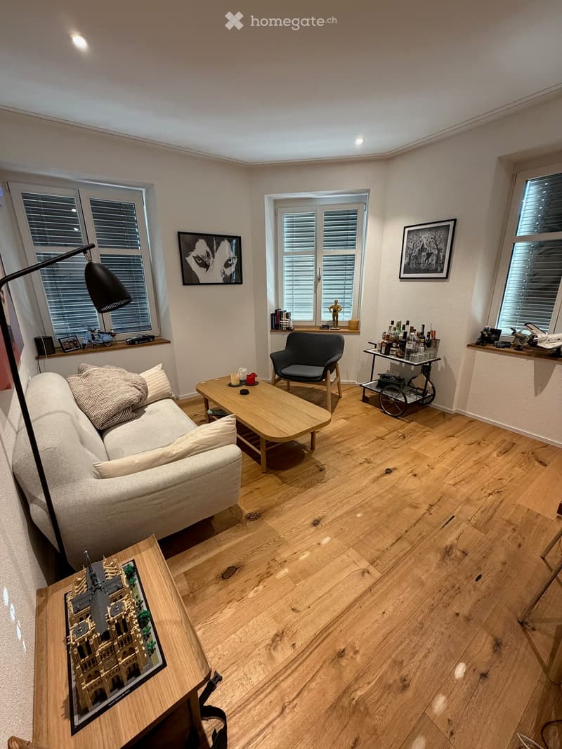 Living room with light wooden flooring, large windows with blinds, a sofa with pillows, a coffee table, a wooden side table with a model display, a bar cart, and a floor lamp.