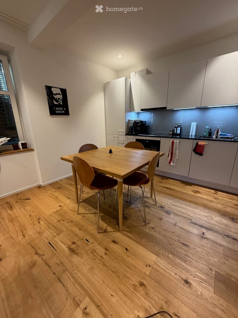 Modern kitchen with light wood flooring, white cabinets, a dining table with four chairs, and various appliances including a refrigerator, oven, and coffee machine.