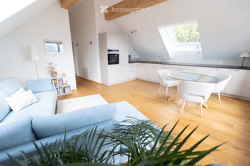 Bright, modern living and dining area with light wooden flooring, white walls, and exposed wooden beams. Features a light blue sofa, white dining chairs around a glass table, a bookshelf, and a small kitchen area with white cabinets and an oven.
