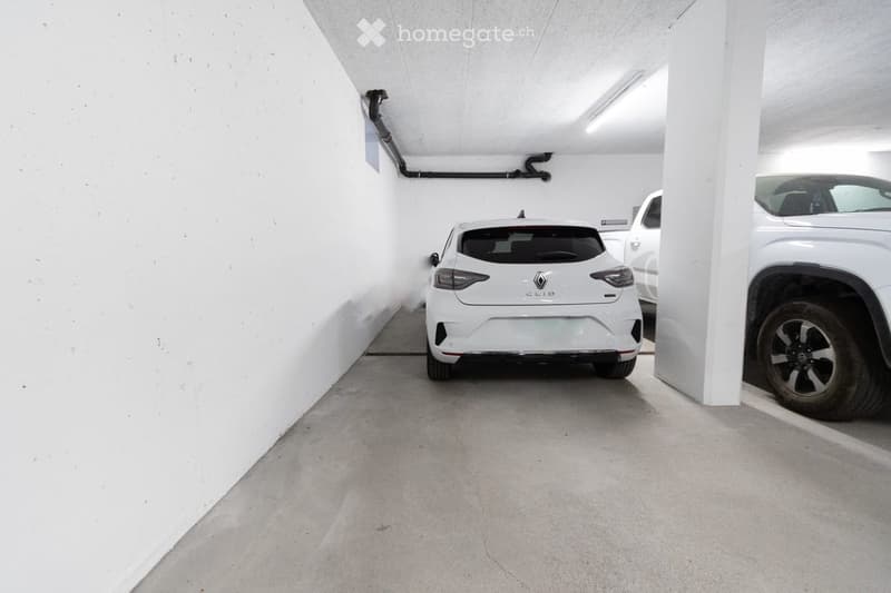 Underground parking area with three cars parked, white walls, concrete floor, and visible structural beams and pipes.