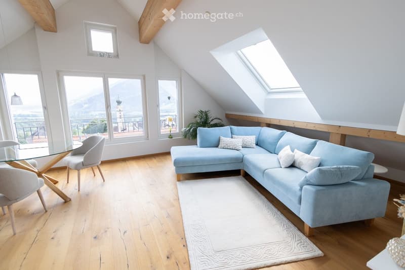 Bright living room with wooden floors, large windows offering a scenic view, a light blue sectional sofa, a glass coffee table, and a white rug.