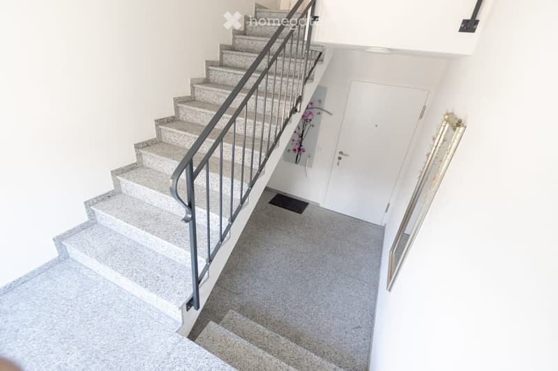 A modern staircase with grey marble steps and a black metal railing leads upwards. The hallway has white walls and a grey carpeted floor, with a closed white door visible on the right side.