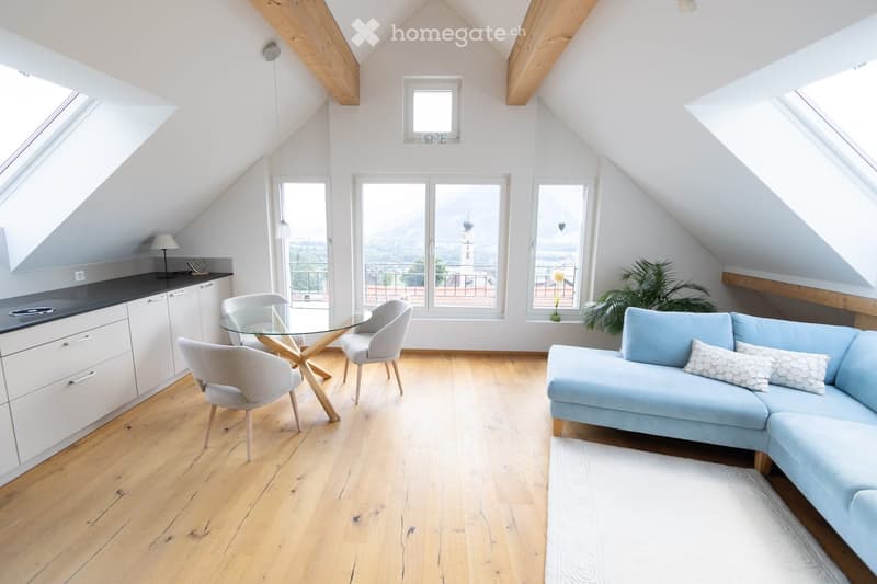 Bright, open loft-style living space with wooden floors, large windows, a modern kitchen area, a glass dining table with chairs, and a light blue sofa.