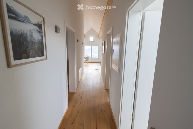 Bright hallway with wooden flooring, white walls, and a large framed landscape painting on the left side. Natural light streams in from the end of the corridor, revealing a glimpse of a room with a chair and a view through a large window.