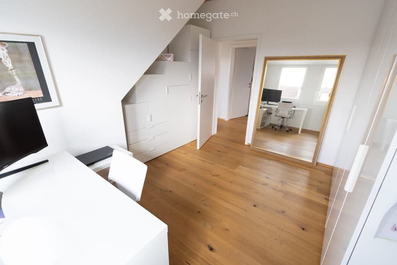 Bright, modern room with wooden flooring, white walls, a large mirror, a desk with a chair, and a cabinet. A TV is visible on the left side.