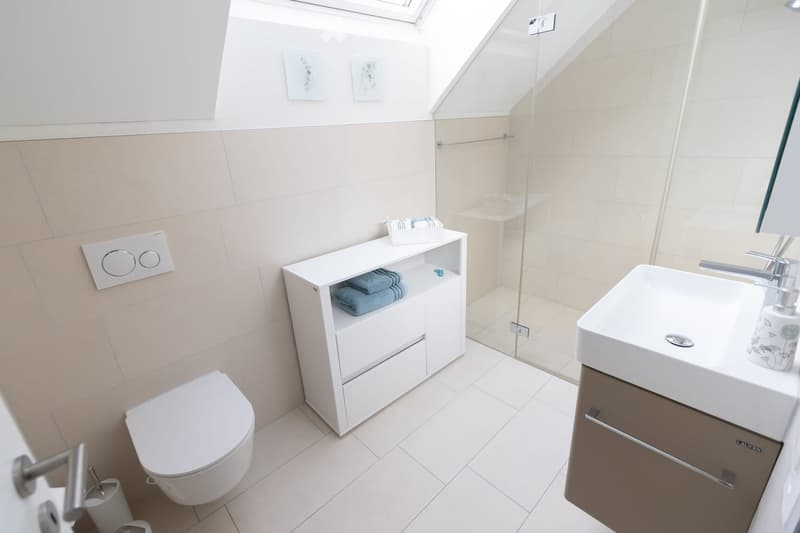 Modern bathroom with white tiles, a toilet, a sink with a mirror, a vanity cabinet with towels, and a glass-enclosed shower area.