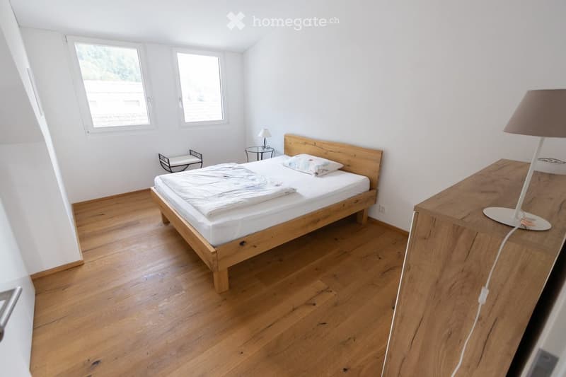 Cozy bedroom with wooden flooring, a single bed with white bedding, a nightstand with a lamp, a small table with a chair, and a window with a view.
