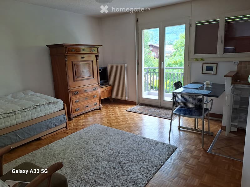 A furnished room with a bed, wardrobe, table, chair, radiator, and a view through large windows.