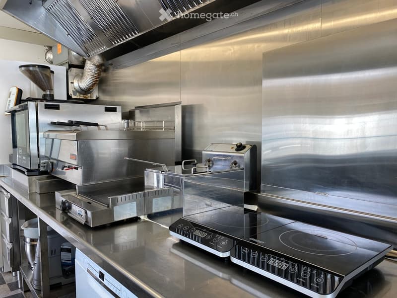 Commercial kitchen with stainless steel surfaces, featuring a coffee machine, induction cooktop, oven, toaster, blender, and refrigerator.