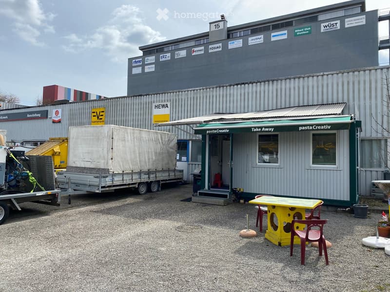 Outdoor area with a small green and white awning labeled "pasta Creativ" and "Take Away" with a yellow table and red chairs, a trailer, and a large industrial building in the background.