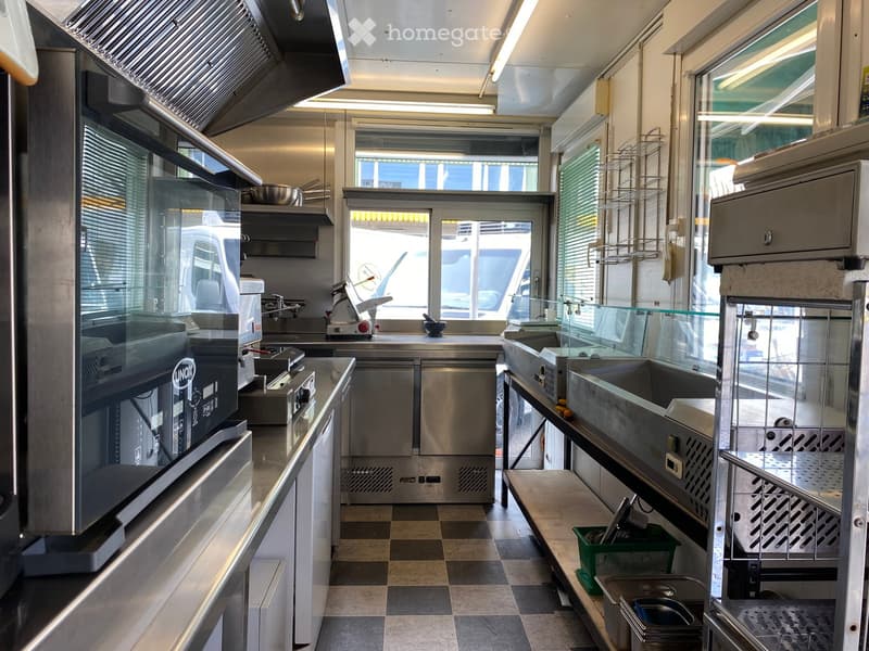 Commercial kitchen with stainless steel appliances, including ovens, a fridge, sinks, and counters. The space is well-lit with large windows and has a clean, organized layout.