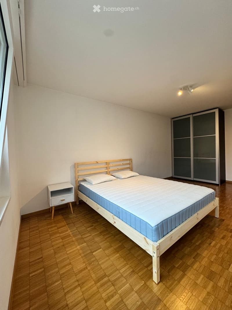 Minimalist bedroom with a wooden bed, blue mattress, white pillows, a small nightstand, and a modern wardrobe with mirrored doors.