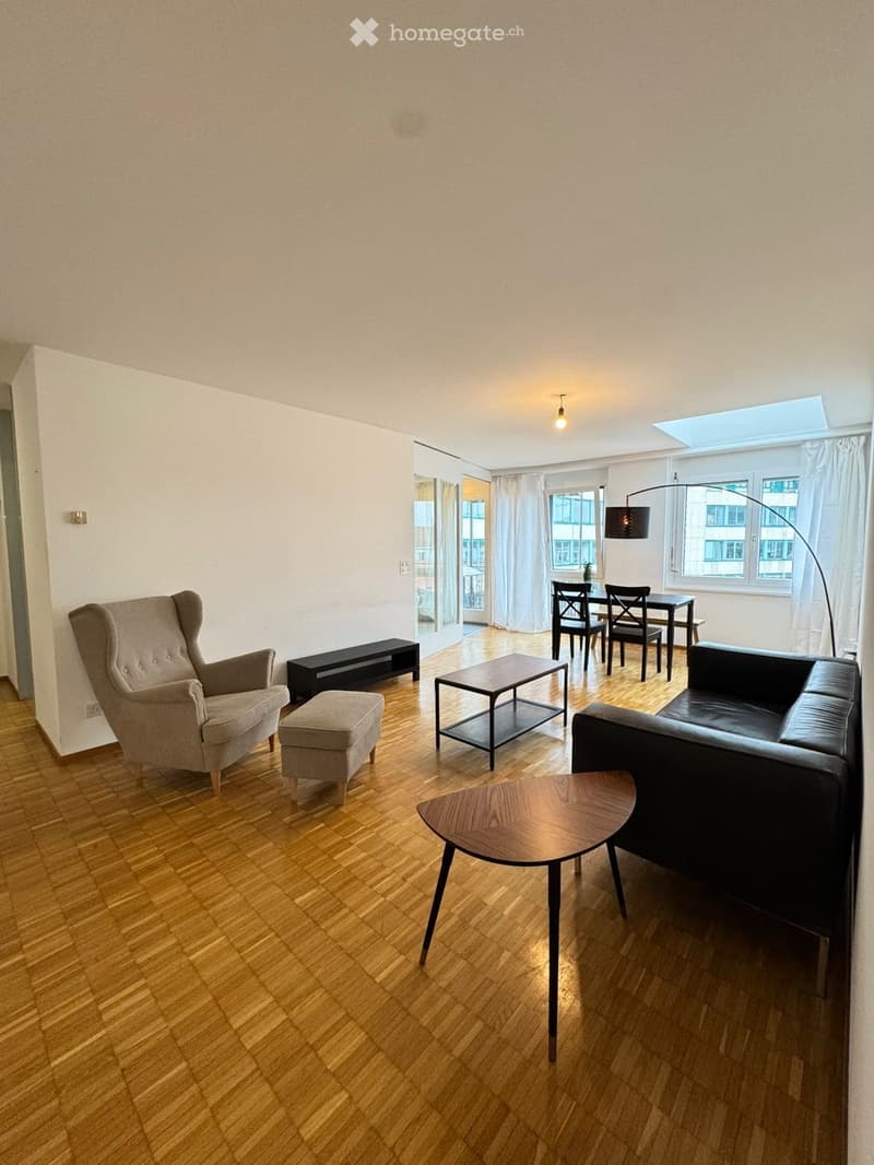 Modern living room with wooden flooring, a grey armchair, a black sofa, a small coffee table, a round wooden side table, a black bench, and a dining area with a table and chairs.
