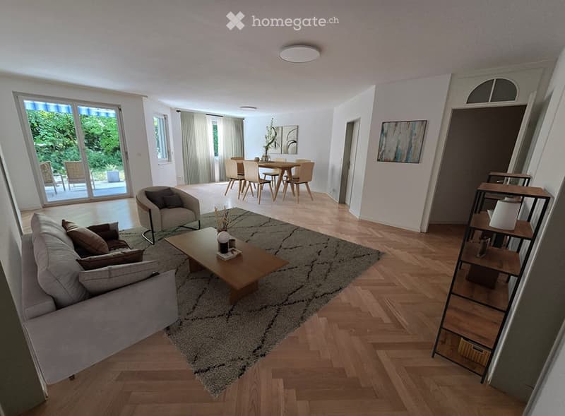 Open-plan living and dining area with hardwood flooring, large windows, a grey sofa, coffee table, armchair, dining table with chairs, and a shelf with decorative items.
