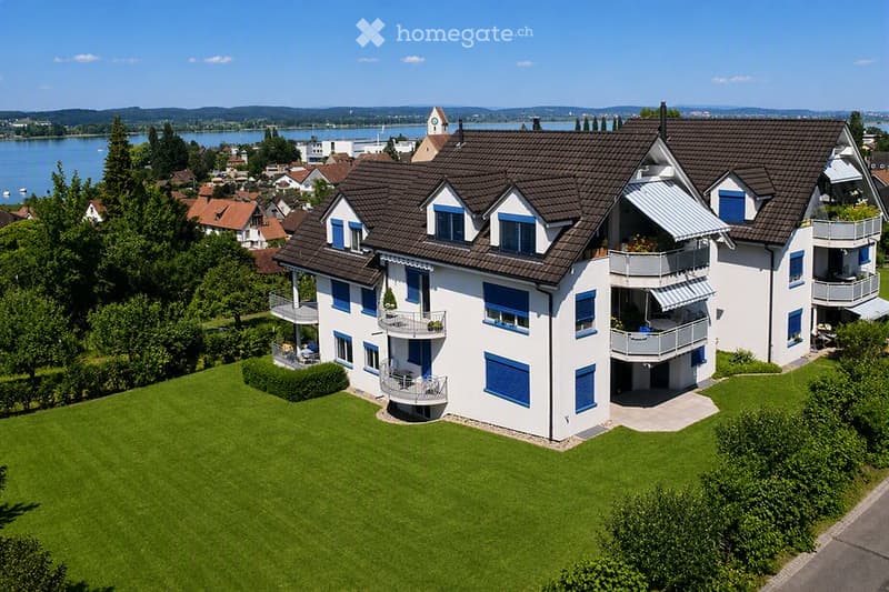 A modern two-story house with white walls, blue shutters, and multiple balconies overlooking a scenic lake and town.