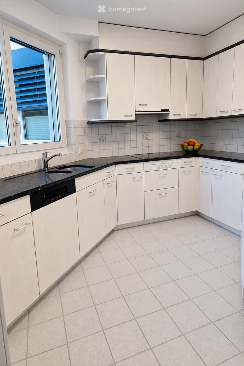 Modern kitchen with white cabinets, black countertops, tiled backsplash, and a window with blinds. Includes a sink, faucet, dishwasher, stove, fridge, and a fruit bowl on the counter.